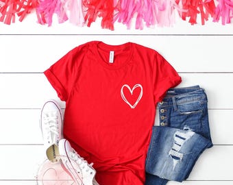 May include: Red t-shirt with a white hand-drawn heart on the front. The shirt is laid flat on a white surface with a pair of white sneakers and blue jeans.