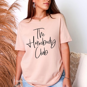 Puede incluir: Una camiseta de color melocot&oacute;n claro con el texto "The Homebody Club" en escritura negra. La camiseta es de cuello redondo y parece estar hecha de un material suave. La marca "C&D's Custom Tees" est&aacute; en la parte inferior.