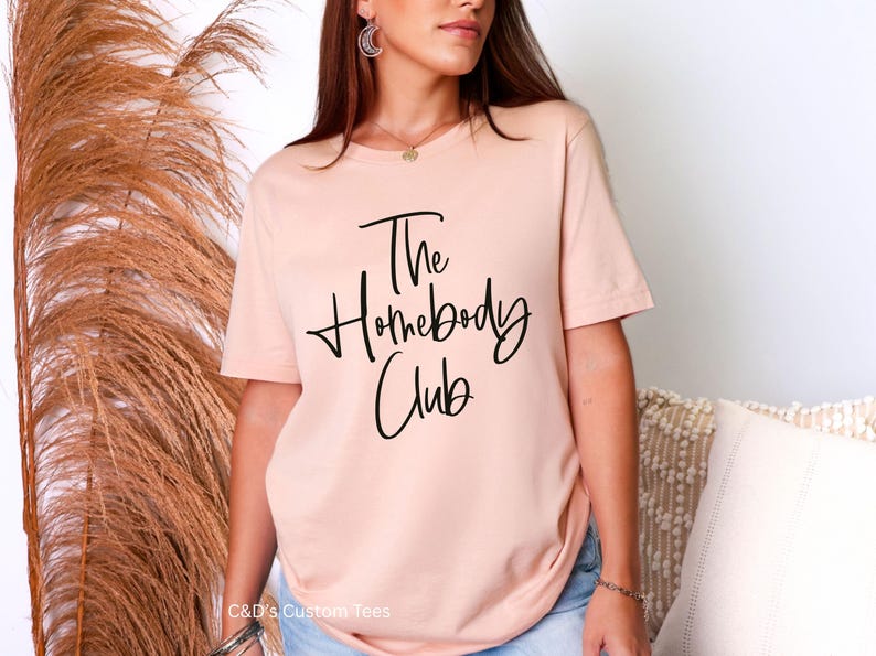 Puede incluir: Una camiseta de color melocot&oacute;n claro con el texto "The Homebody Club" en escritura negra. La camiseta es de cuello redondo y parece estar hecha de un material suave. La marca "C&D's Custom Tees" est&aacute; en la parte inferior.