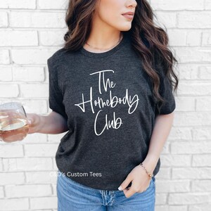 Puede incluir: Camiseta gris oscuro con el texto "The Homebody Club" en escritura blanca. La camiseta se usa con vaqueros azules. Una persona sostiene un vaso de l&iacute;quido.