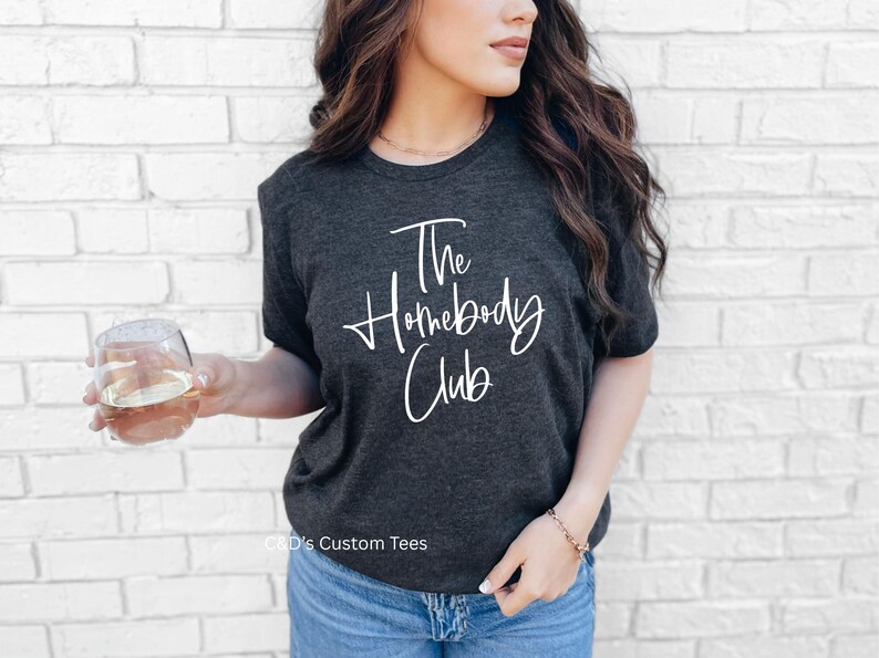 Puede incluir: Camiseta gris oscuro con el texto "The Homebody Club" en escritura blanca. La camiseta se usa con vaqueros azules. Una persona sostiene un vaso de l&iacute;quido.