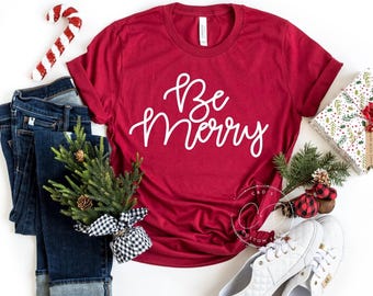 Könnte beinhalten: Rotes T-Shirt mit weißem Text, der "Be Merry" lautet. Das Shirt liegt flach auf einer weißen Oberfläche mit einer blauen Jeans, einem kleinen Kiefernbaum, einem Zuckerstangen und weißen Schuhen.