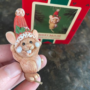 May include: A brown and white mouse wearing a red and white Santa hat with holly and a green stem. The mouse is holding its arms up in the air. The ornament is hanging from a gold string.