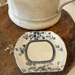 May include: A small, antique, off-white ceramic plate with a blue floral and bird design. The plate has a rectangular center and a scalloped edge. A large, off-white ceramic pitcher is in the background.