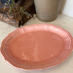 May include: An oval-shaped ceramic serving platter in a solid dusty pink color. The platter features a scalloped edge with a raised, embossed rim detail that follows the contour of the oval shape, creating a smooth and glossy finish throughout.