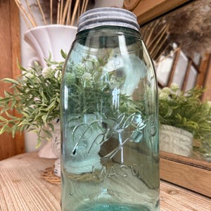 May include: A vintage blue-green Ball Mason jar with a silver lid. The jar has embossed lettering and is filled with greenery. The jar is sitting on a wooden surface, with a white vase and mirror in the background.