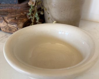May include: A small, round, off-white ceramic bowl with a smooth, glossy finish and a thick, rounded rim. The bowl features a simple, unadorned design with a deep basin and a sturdy base, presenting a clean and neutral appearance.