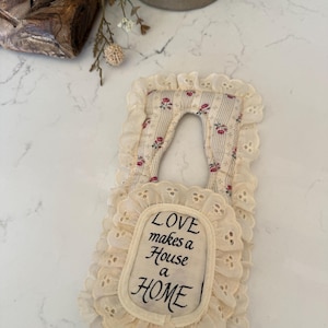 May include: Cream-colored door hanger with a floral pattern and ruffled edges. The text "LOVE makes a House a HOME" is embroidered on the front. A decorative item for the home.