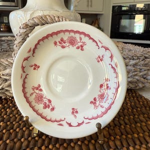 May include: White ceramic saucer with a red floral pattern around the rim and center. The saucer is displayed on a gold-colored stand, showcasing its vintage design. The floral design includes flowers and leaves.
