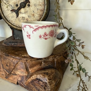 May include: A small, white teacup with a red floral design around the rim and on the side. The cup sits on a carved wooden base, with an antique scale in the background. The scale has a circular face with black numbers and a needle.