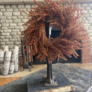 May include: A decorative wreath made of dried, reddish-brown flowers, displayed on a vintage metal stand atop an antique book. The wreath is circular and full, with a natural, rustic appearance. The stand is dark metal, and the book has aged, worn pages.