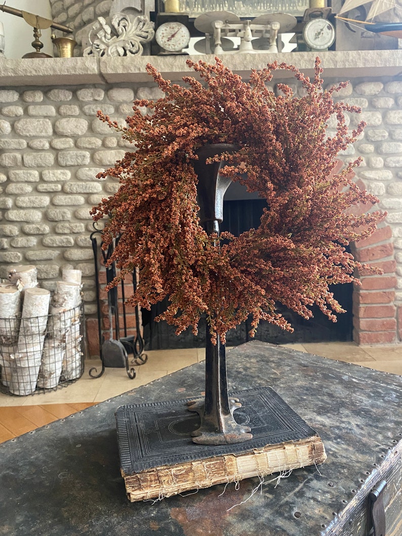 May include: A decorative wreath made of dried, reddish-brown flowers, displayed on a vintage metal stand atop an antique book. The wreath is circular and full, with a natural, rustic appearance. The stand is dark metal, and the book has aged, worn pages.