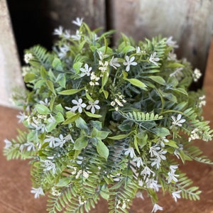 May include: A green and white artificial plant arrangement with small white flowers. The plant is in a white pot and is sitting on a wooden surface.