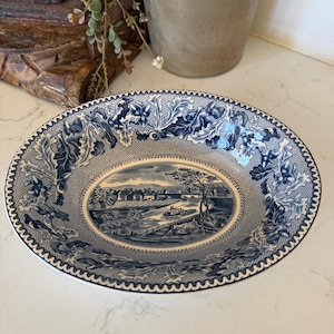 May include: An oval ceramic bowl featuring a blue and white transferware pattern. The center depicts a pastoral landscape with a canal and buildings, while the rim is decorated with a detailed border of oak leaves and acorns on a mesh background.