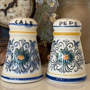 May include: A pair of cream-colored ceramic salt and pepper shakers featuring hand-painted blue and yellow floral patterns. The left shaker is labeled SALE in black text, and the right shaker is labeled PEPE in black text, both with blue rims.