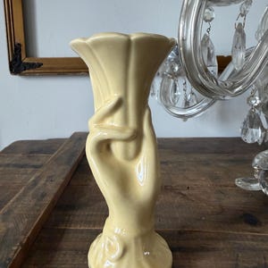 May include: A pale yellow ceramic vase in the shape of a hand holding a flower. The vase has a flared top and a round base. The hand is detailed with curved lines and a smooth finish. The vase is likely vintage or antique.