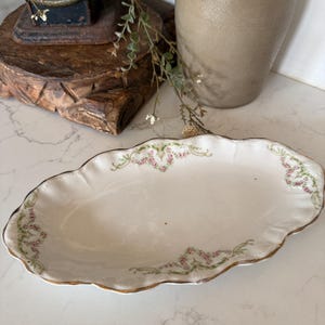 May include: An oval porcelain serving dish with a scalloped edge and a gold-trimmed rim. The white surface features delicate floral garlands in pink and green, arranged in a symmetrical pattern along the curved border of the dish.