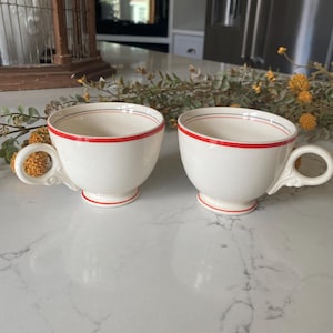 May include: Two white ceramic teacups with a red and blue trim. The cups have a classic design and are perfect for a tea party.