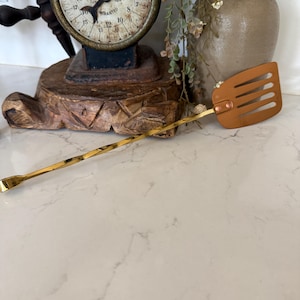 May include: A gold-toned metal kitchen utensil featuring a long, twisted handle and a flat, slotted rectangular head. The head is attached to the handle with a small copper-colored rivet, and the opposite end of the handle features a small, curved scoop.
