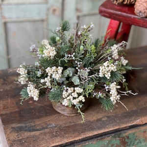 May include: A small white berry and green foliage arrangement in a rustic brown pot. The arrangement is sitting on a wooden surface with green paint.