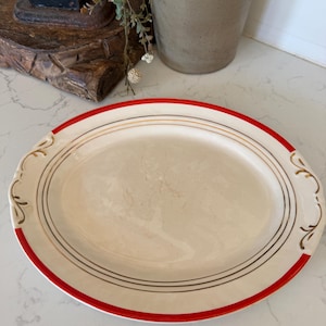 May include: An oval ceramic serving platter featuring a cream-colored center with thin black and gold circular bands. The rim is accented with bold red sections and decorative gold scrollwork details on the left and right sides of the plate.