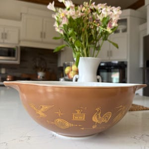 May include: A brown Pyrex mixing bowl with a white interior and gold-colored farm-themed graphics. The bowl features a rooster, eagle, and other vintage designs. It has two handles and is set on a white countertop.