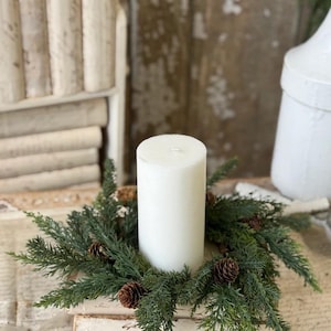 May include: A white pillar candle sits in the center of a circular wreath made of green faux pine needles and small brown pinecones. The candle and wreath are positioned on top of a stack of two off-white books with visible text on the covers.