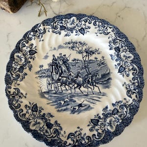 May include: A round ceramic plate with a scalloped edge featuring a blue and white transferware pattern. The center depicts a horse-drawn carriage with passengers and running dogs. The border is decorated with floral motifs and foliage.