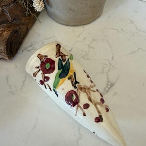May include: A cone-shaped ceramic wall pocket vase with a cream-colored glazed finish. The surface is decorated with hand-painted brown branches, dark red berries, and a small green and yellow bird perched among the foliage.