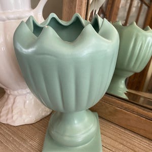 May include: A small, light green ceramic vase with a fluted design and a square base. The vase has a scalloped rim and is perfect for displaying small flowers or greenery.