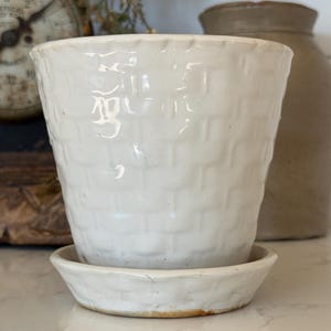 May include: A white ceramic plant pot with a raised basket-weave texture on the exterior surface. The pot features a matching circular saucer base with a slightly uneven, rustic rim and a glossy finish that reflects light across the textured pattern.