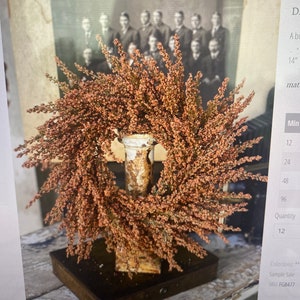 May include: A decorative wreath with small, reddish-brown flowers, mounted on a distressed metal stand. The stand is set on a dark wooden base. A vintage black and white photograph is in the background.