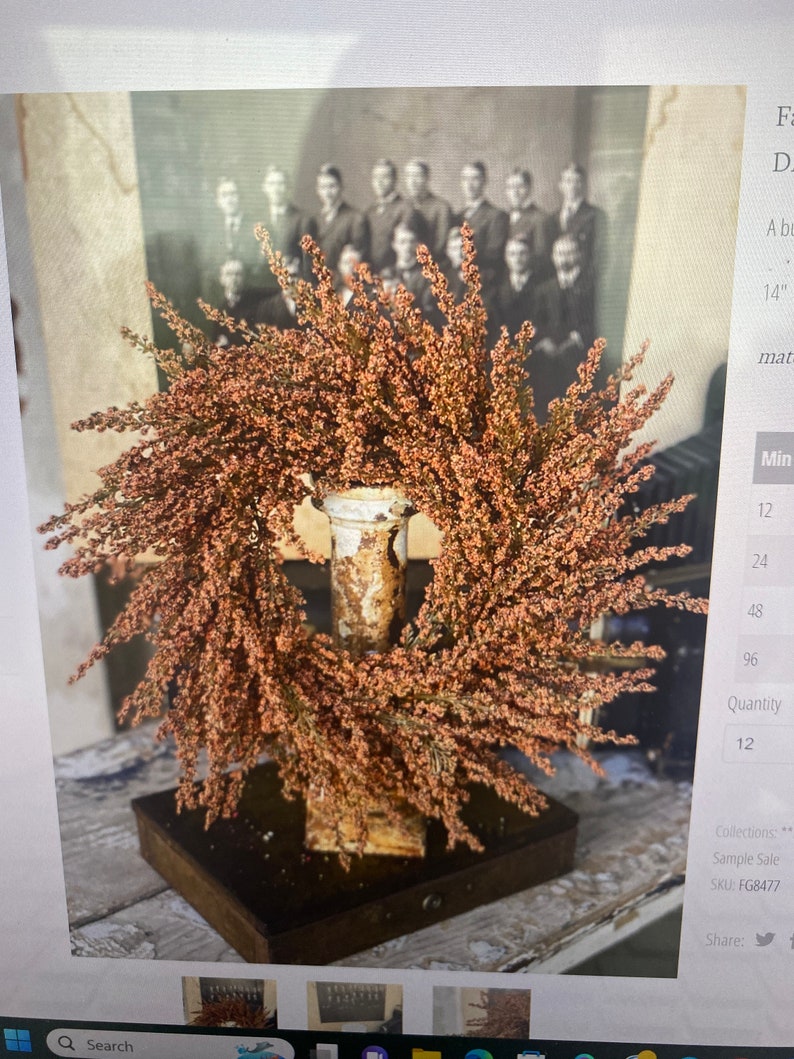 May include: A decorative wreath with small, reddish-brown flowers, mounted on a distressed metal stand. The stand is set on a dark wooden base. A vintage black and white photograph is in the background.