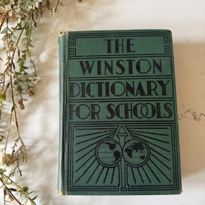 May include: A vintage green hardcover book titled The Winston Dictionary for Schools. The front cover features black embossed text and a graphic illustration of two globes with radiating lines. The book has a rectangular shape and a worn spine.