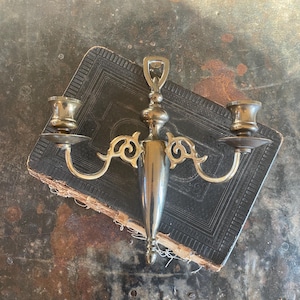 May include: A gold-toned metal wall-mounted candle holder with two candle arms and an ornate design. The holder has a decorative finial at the top and is mounted on a black book.