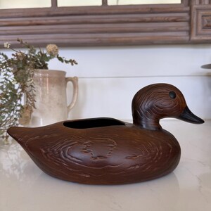 May include: A brown duck-shaped planter with a textured wood grain finish. The planter has a black interior and a black beak. The duck is resting on a white surface. Decorative home decor.