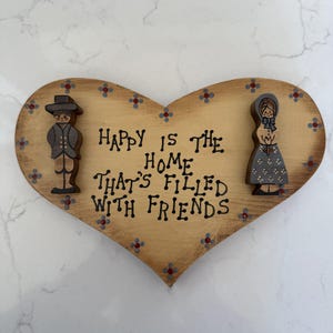 May include: Wooden heart-shaped sign with the text "Happy is the home that's filled with friends." The sign has a light brown background with painted floral accents and two wooden figures on either side.