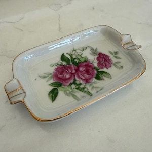 May include: A rectangular, white ceramic ashtray with a floral design featuring pink roses and green leaves. The ashtray has gold trim and small handles on each end.