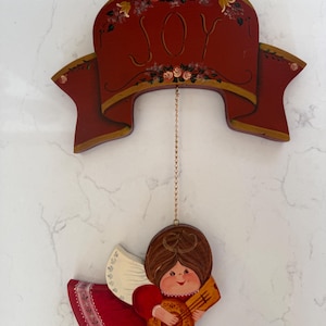 May include: A wooden Christmas ornament featuring a red banner with the word "JOY" and floral accents. Below, a painted angel in a red dress plays a guitar. The ornament hangs from a gold chain, perfect for holiday decor.