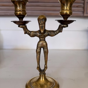 May include: A vintage brass candlestick holder featuring a stylized figure with outstretched arms holding two candle cups. The figure has a beard and is standing on a round base. The candlestick holder is a warm gold color.