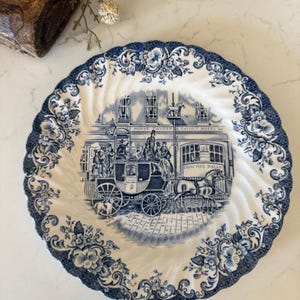 May include: A round ceramic plate with a scalloped edge featuring a blue and white transferware pattern. The center depicts a horse-drawn carriage in front of the White Horse Tavern & Family Hotel with text reading Coaches Daily.