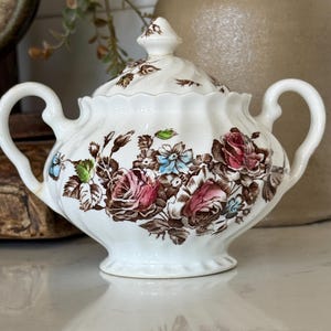 May include: A white ceramic sugar bowl with a matching lid, featuring two curved handles and a fluted rim. The body is decorated with a transferware pattern of pink roses, brown leaves, and small blue flowers. The lid has a pointed finial.