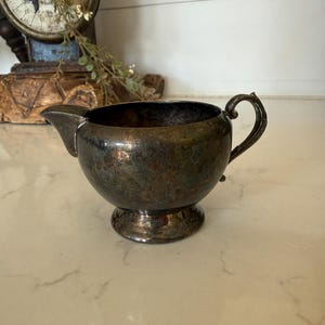 May include: Antique silver creamer with a spout and handle. The creamer has a round body and sits on a small, decorative base. The silver has a tarnished appearance, giving it a vintage look. A classic piece for serving cream or milk.