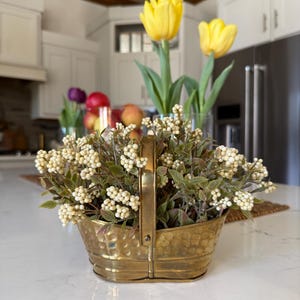 May include: A decorative brass basket filled with white berry stems and green leaves. Two yellow tulips in a glass vase are behind the basket. The basket has a handle and a hammered texture. The background includes white cabinets and a stainless steel refrigerator.