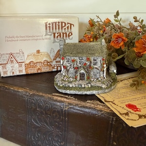 May include: A miniature cottage with a stone facade, adorned with red roses and green foliage. The cottage sits on a base of greenery, next to a box labeled "Lilliput Lane" and a vintage book. The cottage is a handmade miniature.