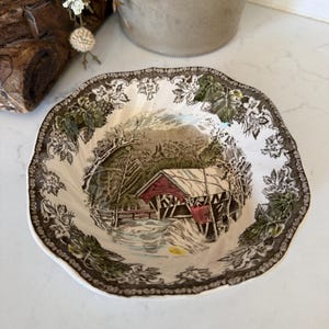 May include: A scalloped ceramic bowl featuring a central illustration of a red covered bridge in a snowy landscape. The rim is decorated with a detailed brown and green botanical pattern of leaves and flowers on a cream-colored background.