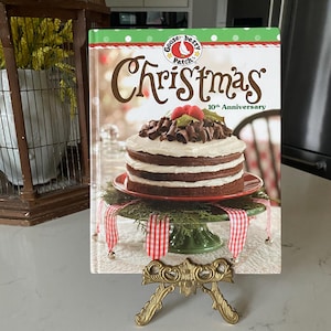 May include: A cookbook titled "Christmas 10th Anniversary" is displayed on a decorative gold stand. The cover features a layered chocolate cake with white frosting, chocolate shavings, and berries. The book is on a green cake stand with red and white ribbon.