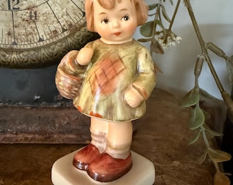 May include: A vintage porcelain figurine of a young girl holding a basket. The girl wears a green dress with a brown patch, brown shoes, and a red bow in her hair. The figurine is on a square base.