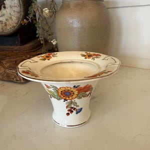 May include: A cream-colored ceramic bowl with a wide, flared rim and two small handles. The exterior and rim feature a colorful floral pattern with orange, yellow, green, and blue blossoms and leaves, accented by a thin gold-toned trim along the edges.