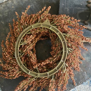 May include: A decorative wreath featuring a circular metal frame with a ring of dried, brown-toned floral elements. The wreath is set against a dark, textured surface, suggesting a rustic or vintage aesthetic. The overall design evokes a natural, autumnal theme.